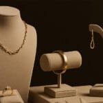 Displays gold jewelry including a necklace, earrings, a bangle and a ring, all placed on beige jewelry stands against a dark background.